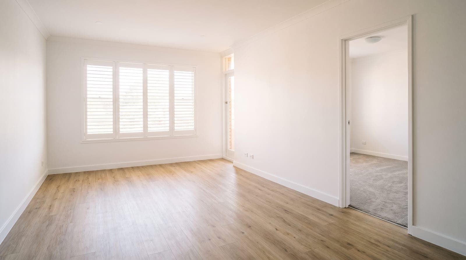 An empty rental living room with timber-look hybrid flooring, opening through a doorway to a carpeted bedroom