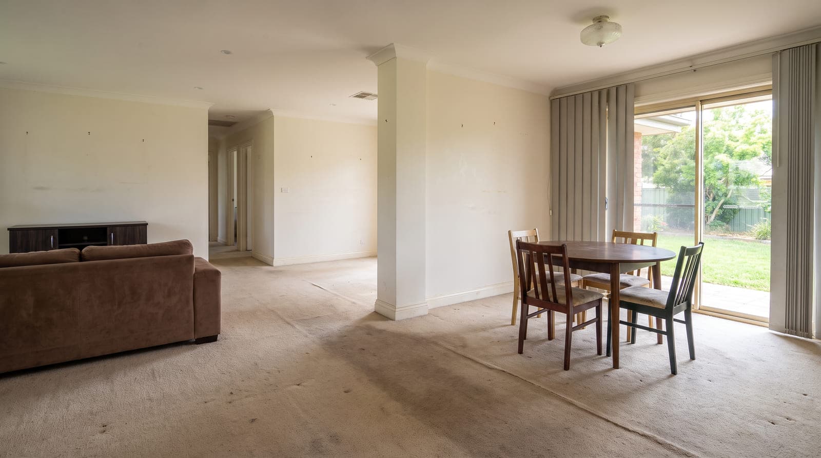 Open living and dining area before renovation — worn beige carpet with a darkened traffic path, dated vertical blinds and tired furniture