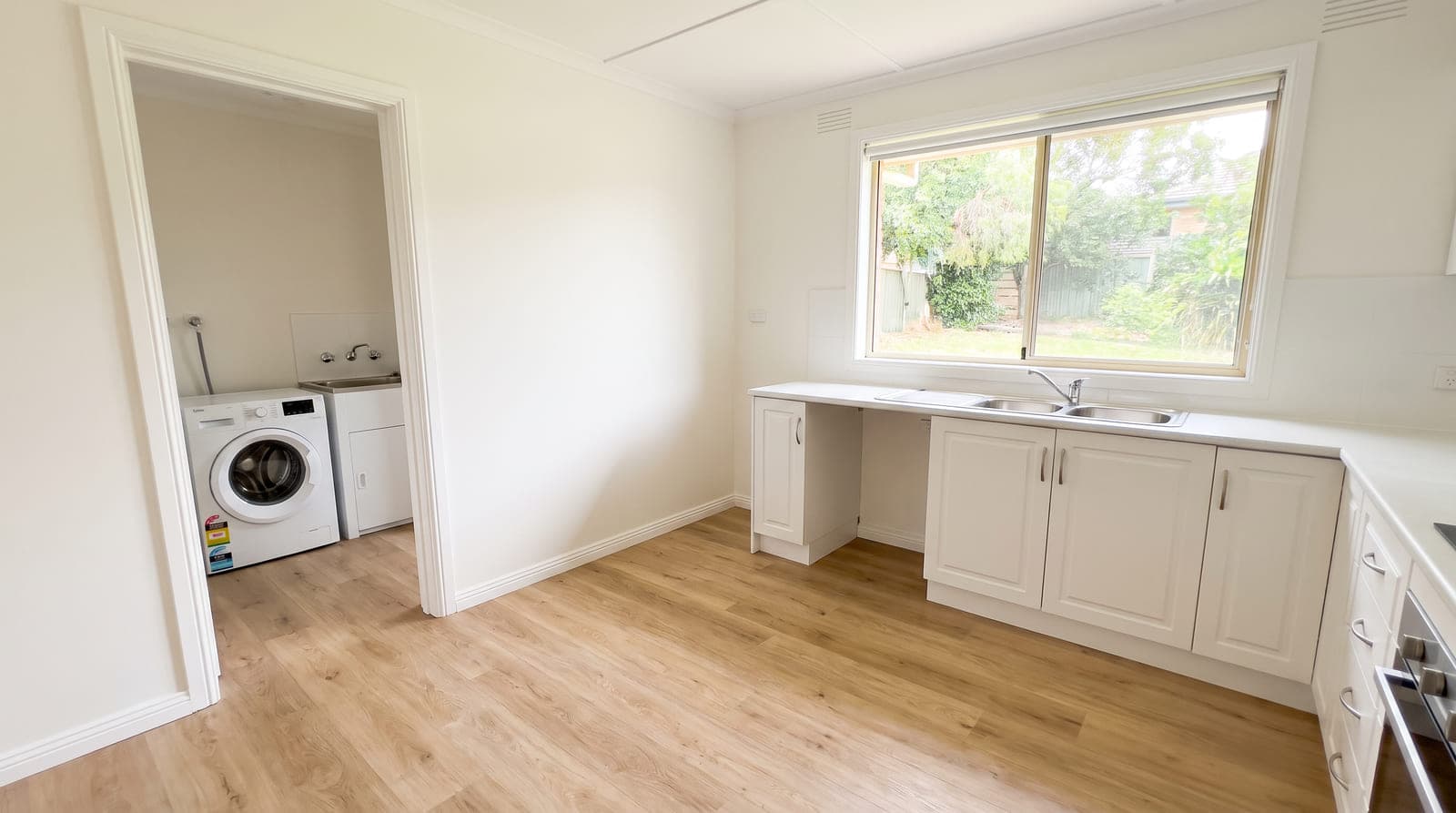 The same kitchen and laundry nook after renovation, with pale oak timber-look vinyl plank running through both rooms without a threshold