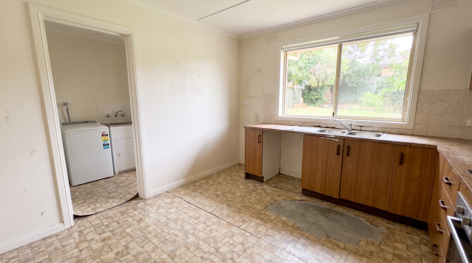 Kitchen and laundry nook before renovation — old patterned sheet vinyl worn through in front of the sink and curling at the doorway, with dated brown laminate cabinetry